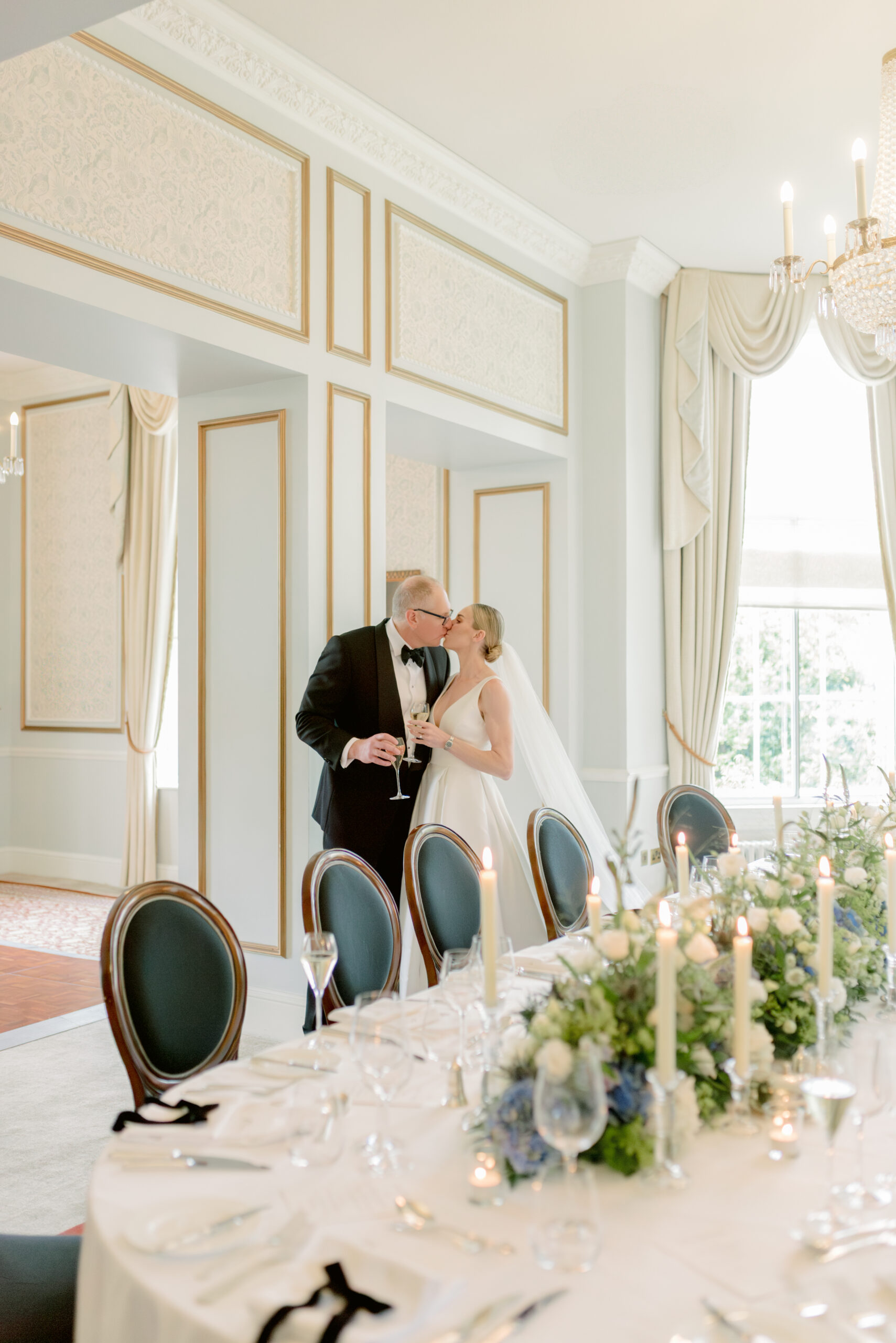 Bride and Groom kiss in the wedding dining room of Gleneagles on their wedding day. With blue, white and green hydrangea table design; floral meadow, velvet bow place settings and taper candles. Image by award winning fine art wedding photographer Scotland, Jill Cherry Porter.