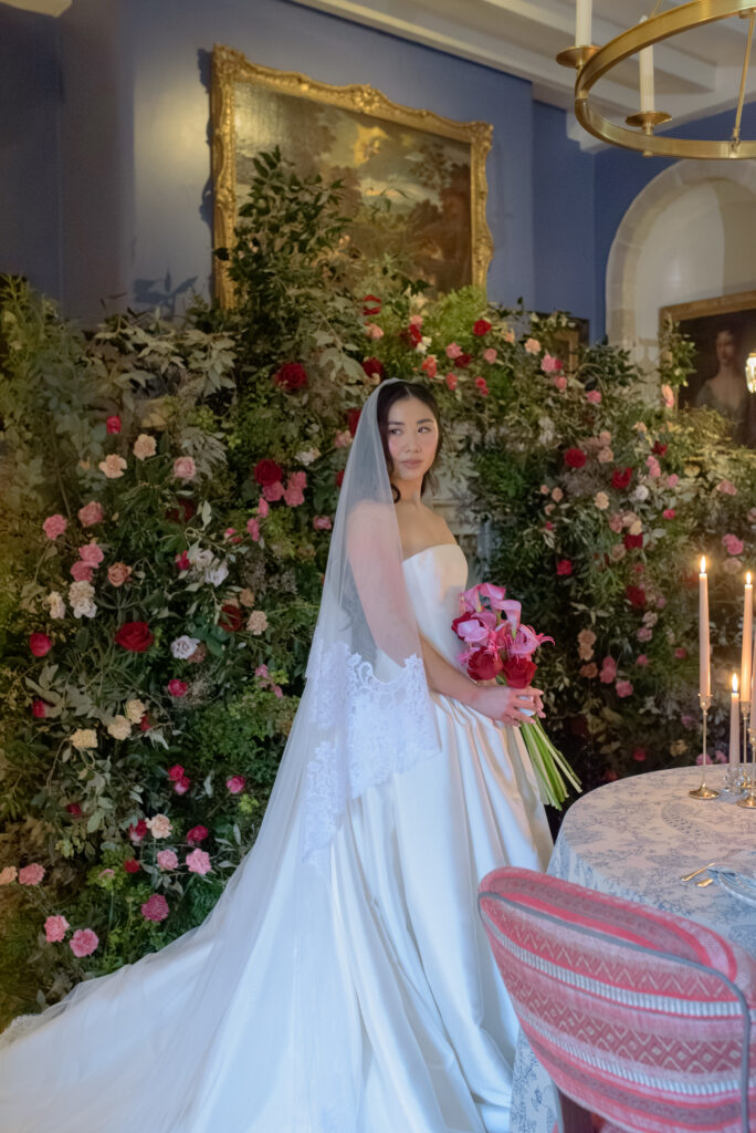 Bride in Emma Beaumont couture wedding gown holding bouquet in front of abundant floral installation at Penicuik House Scotland
